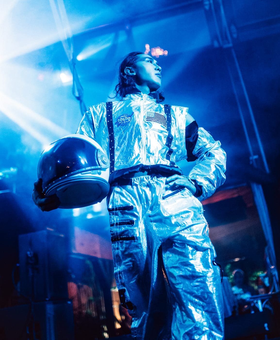 A performer in a silver spacesuit stands under blue light with their domed helmet tucked under one arm