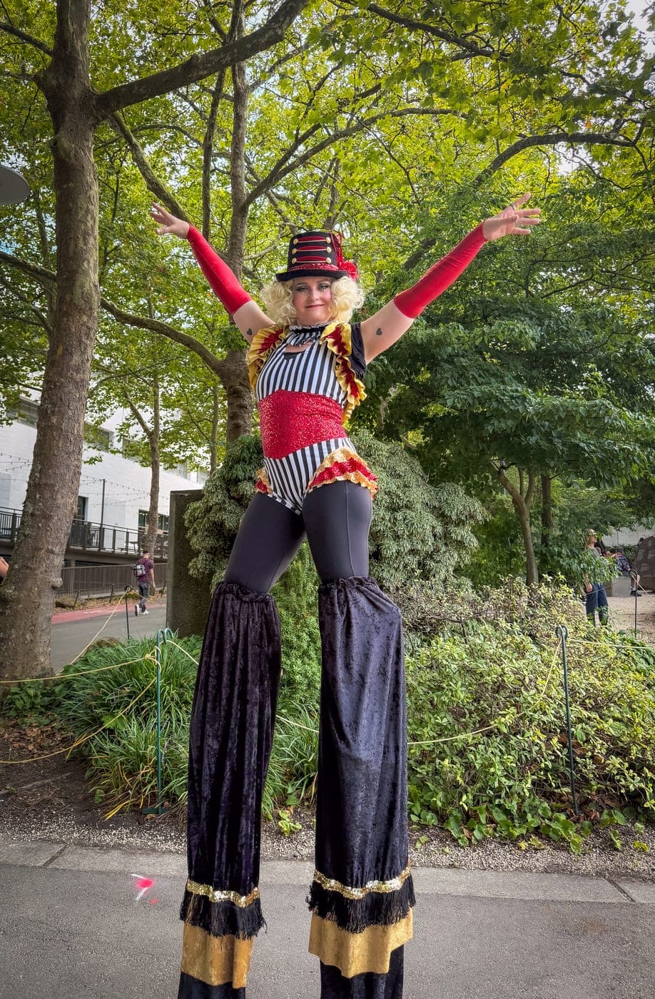 A stilt walker in costume with arms raised, outdoors among trees