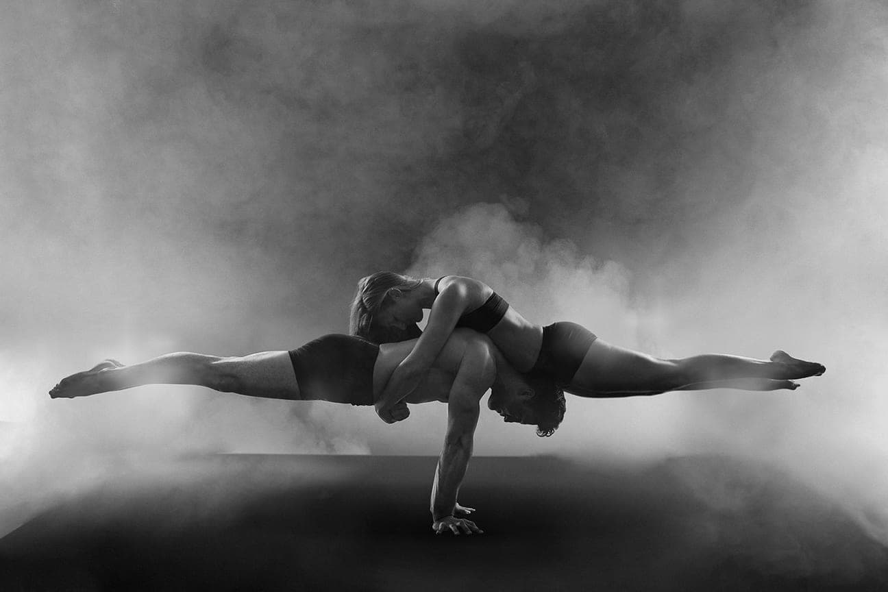 Two acrobats balancing together in a smoke-filled studio, in black and white