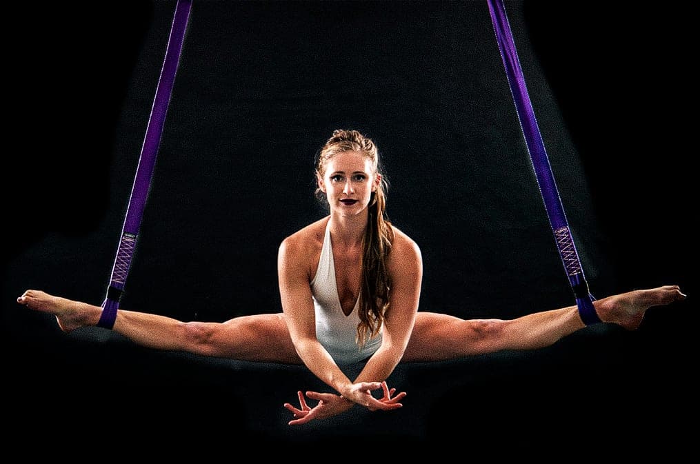 Katie holding a straddle in aerial straps against a dark stage