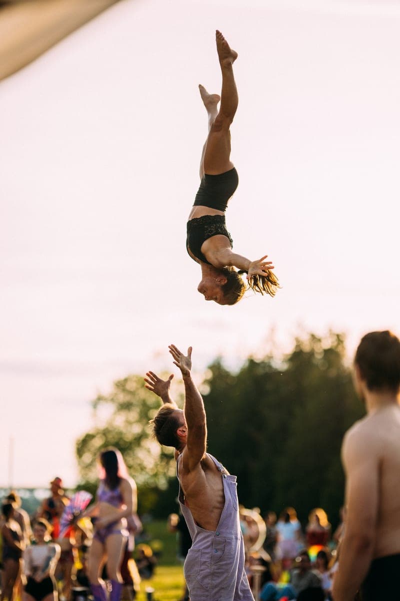 An acrobat thrown high above an outdoor crowd, arms reaching